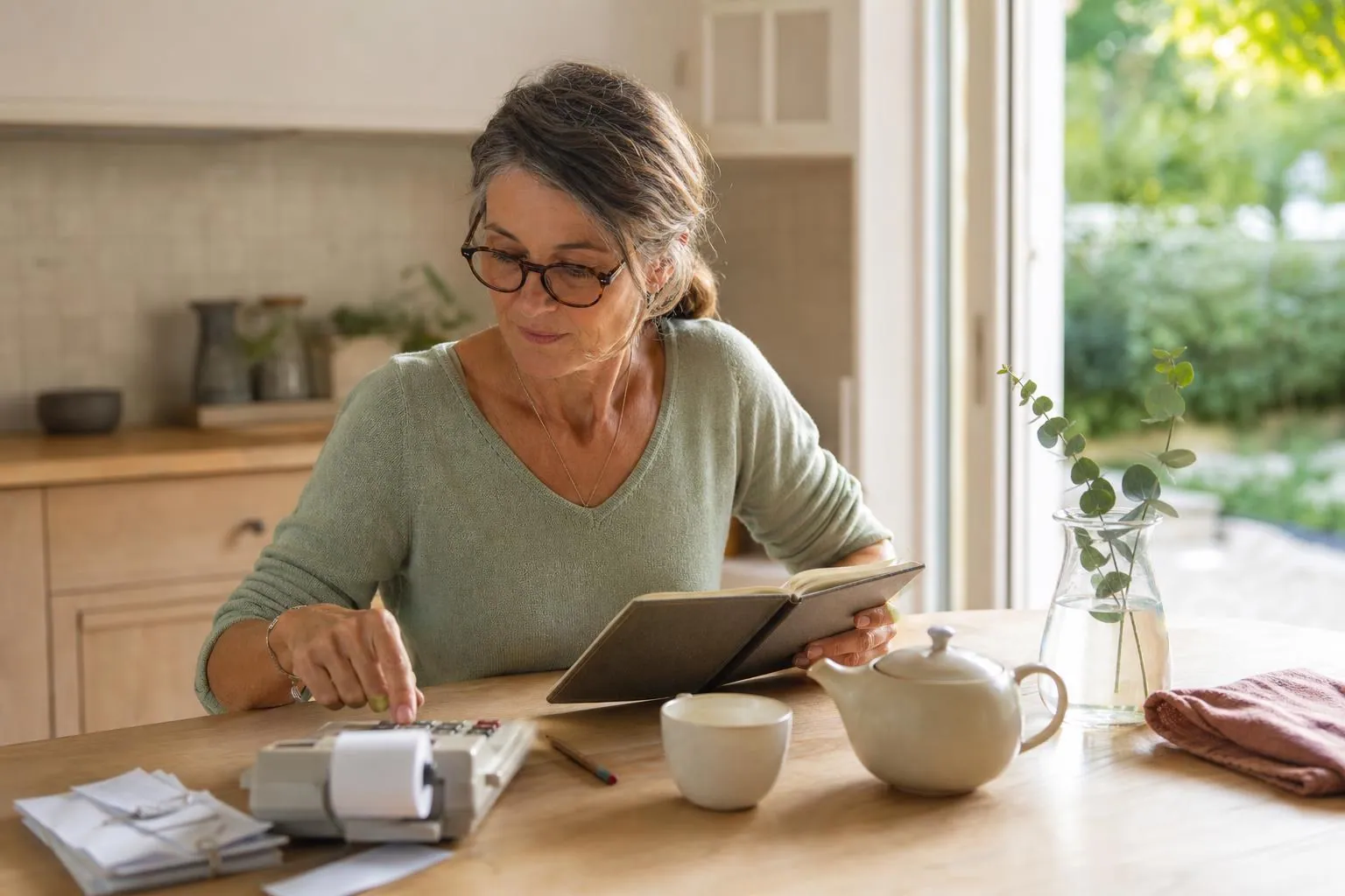 Femme d’une cinquantaine d’années assise à une table de cuisine claire, calculatrice de bureau et carnet de comptes devant elle