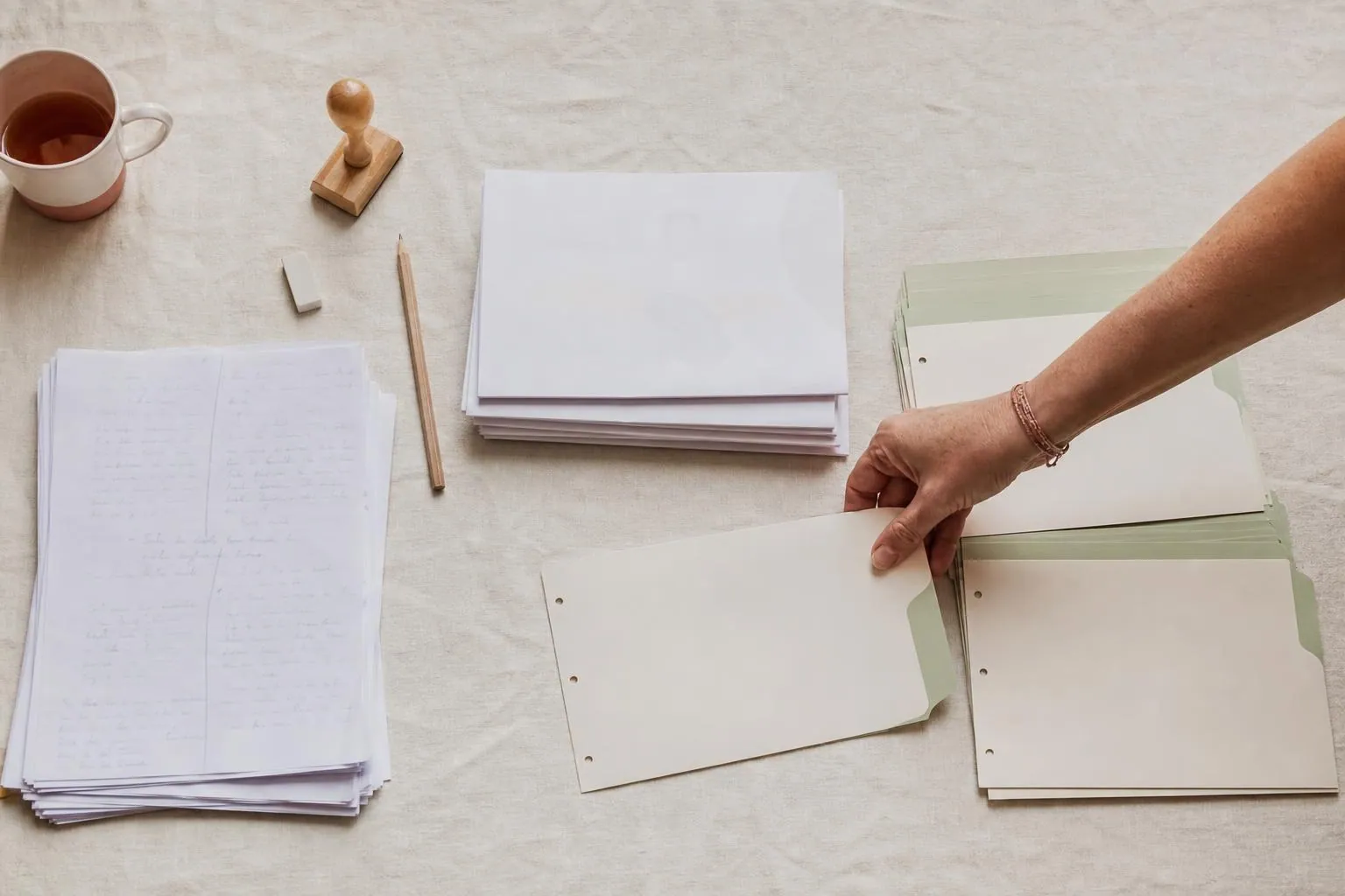 Vue en plongée sur trois piles de documents distinctes posées sur une nappe de lin crème, une main féminine déplaçant une fiche